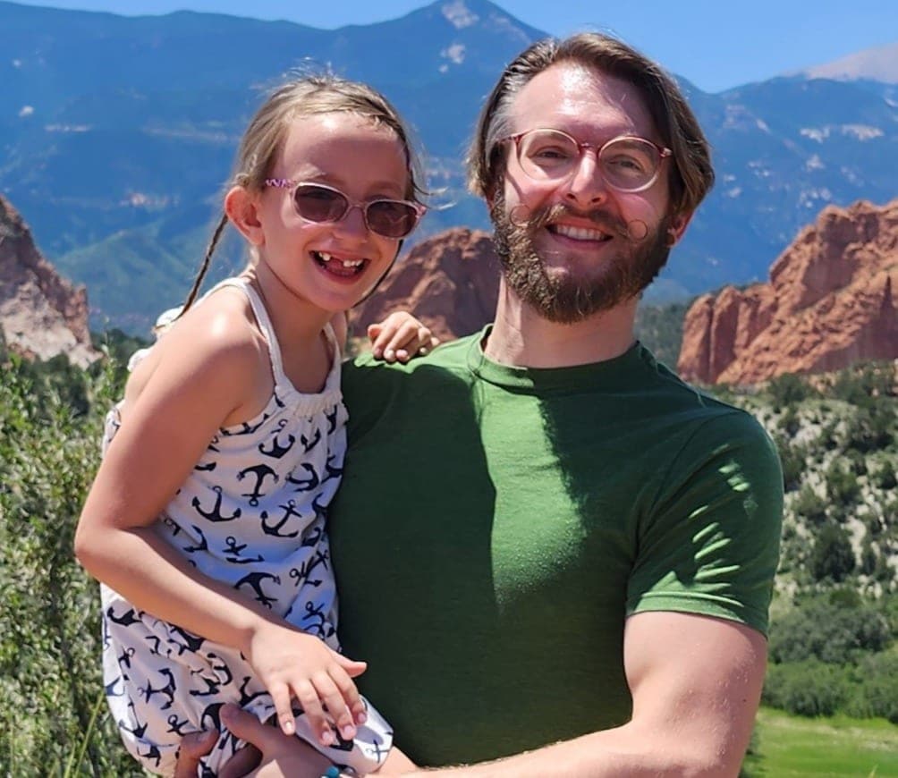 Josh Rafail holding his daughter, with the red rocks and Pikes Peak behind them